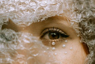 A close-up shot of a young woman with a bubble-wrapped face. One brown-coloured eye stares out from behind a tear in the wrap. There are tiny beads of pearls under her eye that could depict tears.