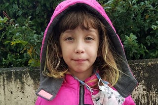 7-year-old kid in a pink hoodie with a mask around her neck holds her new Covid-19 vaccine card and wears a vaccinated sticker