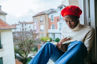 A photo of a young black woman reading by her window.