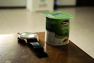 A single serve cup of yogurt next to a wristwatch.