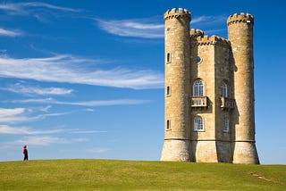 A small medieval-looking tower stands on a grassy hill, with wispy clouds moving on the sky behind it. A solitary woman stands off to the side, looking up at the tower.