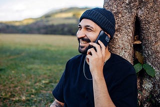 A bearded man in a black shirt and beanie sits in a field with his back to a tree, smiling and talking on a cell phone