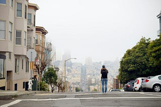 A person taking a photo of a San Francisco street at the top of its hill on a foggy day.