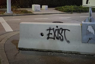 A piece of graffiti on concrete. It reads: “Exist”