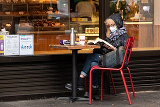 Older woman wearing face mask reading a book outside a cafe.