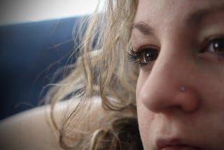 A closeup of a woman’s face. She has a nose piercing and brown eyes and wavy blonde hair.