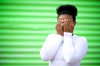 A girl covers her face with her hands against a bright green background.