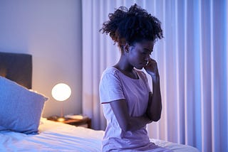 A troubled black woman sits at the edge of her bed.