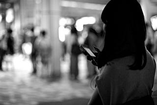 A black and white photo of a black-haired woman on her phone in a busy city street.