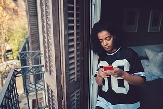 A photo of a black woman on her phone looking out of her apartment window/door.