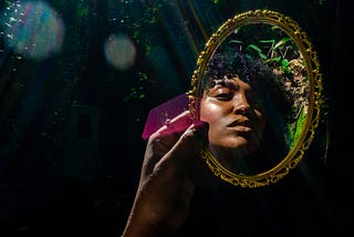 Young Black woman holding up and looking into a gilded mirror in the forest.