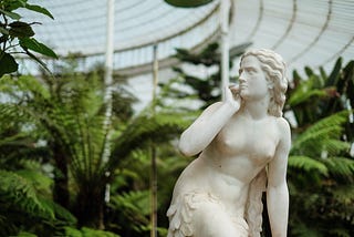 A marble statue of a thin woman looking off to the side, angrily and mistrustfully. The statue stands in front of lush ferns.