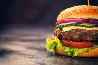 Close-up of a cheeseburger in a sesame seed bun with lettuce and sliced onions, tomatoes, and cucumbers on a black background