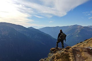 A man with a backpack stands on a trail overlooking remote mountains.