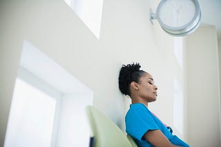 A photo of a tired black female nurse sitting in a hospital hallway chair.
