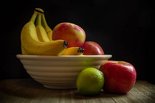 A still-life photograph of a bowl of fruit, with bananas, red apples, and a lime