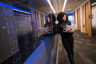 A woman looks at her laptop while leaning on the glass in front of a row of computer servers