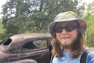 The author is wearing a tie-die shirt and hat. His beard and mustache are beginning to turn white. He’s wearing sunglasses and is standing in front of a rusty abandoned vintage car in the middle of the woods.