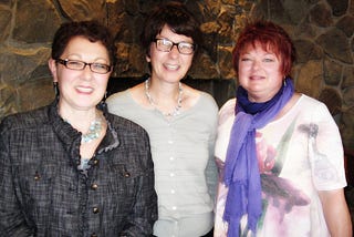 Photo of three women in front of a stone wall, smiling and facing the camera.