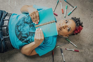 Black woman lying on the floor holding a notebook and smirking, surrounded by pens.