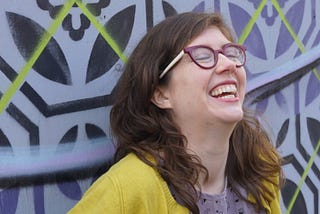 Woman laughing and smiling with her head tilted back again a wall covered with a geometric mural.