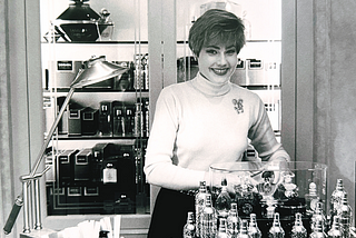 Woman with dark short hair, a large smile, at a perfume counter. Image of author at age 35, selling Guerlain fragrances in Bergdorf Goodman.