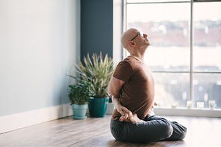 Photo of me seated in lotus pose, holding my feet. My head is tilted skyward. I am next to a window overlooking downtown Pittsburgh.