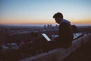 man on a ledge with his laptop at the rooftop overlooking the city sunrise