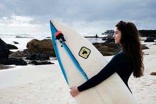 three australian accent variations girl surfer holding surfboard on beach looking out at water sand and rocks smiling