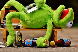 Kermit The Frog lying on wooden bench surrounded by glass bottles. Kermit appears drunk with a bottle of clear bottle presumed to be vodka in one hand and a brown bottle presumed to be beer in the other. He’s lying on his back with his mouth open and one leg hanging over the back of the bench.