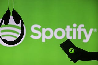 Headphones hang upside down as a hand holds a phone with the Spotify logo displayed in front of a screen showing Spotify
