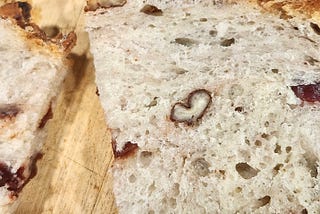 Color square closeup image of one of the pieces of homemade walnut cranberry wheat bread from the prior image. Where we see actually two cut slices of the bread laying on a light wooden cutting board, and the camera eye is trained on the slice to the right of viewer showing how this bread piece sliced caused in it’s top middle section of the slice to have one of the walut pieces sliced through look like the outline of a small ‘heart’.