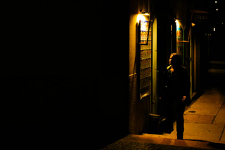 A person outside of a bar at night, illuminated by warm light, the other half of the frame cast in shadows.
