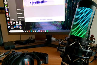 A HyperX microphone and headset on a desk, in front of a monitor showing an open audio editing program. The microphone is lit in green and blue.