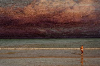 A photo of a man walking on the beach. The sky is dark with red clouds.