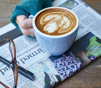A hand holding a cup of coffee over a newspaper with a pair of glasses and a pen.