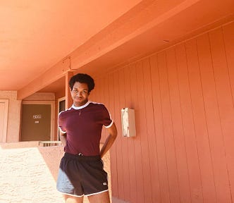 A young Black man with short natural hair, dark skin, and dark eyes, wearing a maroon colored t-shirt and black shorts stands in front of an orange-painted building with his hands on his hips.