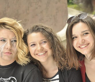 Three young 20-something, light-skinned, women/femme presenting humans. On the left — short blonde hair, gray and white face paint on cheeks and faded rainbow painted on eyelids, not smiling, bangs hanging down on one side, black shirt; Middle — brown, medium-length, wavy hair, smiling, black and white striped shirt with black sweater; On the right — brown, straight, medium-length hair, smiling, sunglasses on head, black top with red sweater