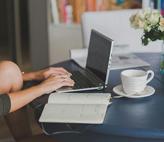 A white woman is sitting at a desk, typing on her laptop. A mug, journal, and vase of flowers are on the table too.