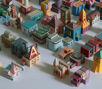 A picture of Charles Young’s paperwork: There are dozens of tiny buildings and vehicles made from colorful paper arrayed together on a white table, so that it looks like a city seen from above