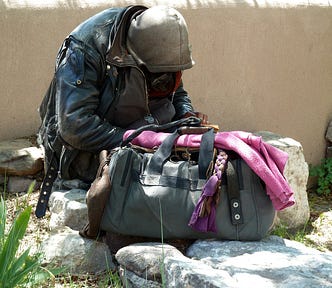 A person hunched over wearing a worn leather jacket and dirty hoodie, sitting on large rocks against a wall with a grey duffel bag, pink sweater on top.