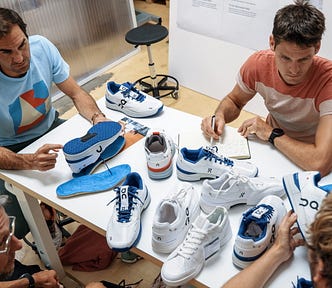 Roger Federer participates in a design meeting with On running shoe developers, gathered around a wooden table covered with prototypes of white and blue athletic footwear. Federer points to an insole while another designer takes notes. Several different color variations of the shoes are visible, including one with red and orange accents. The scene suggests a collaborative process focused on refining the design and performance of the On tennis shoe.