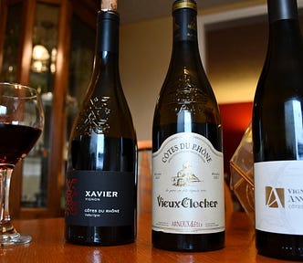 The picture shows three wine bottles from France. There is a wine glass on the table half full of red wine.