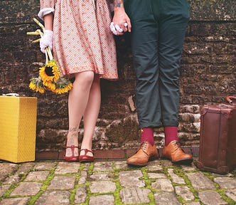 Light-skinned woman holding a bunch of sunflowers, wearing a red and white dress, white gloves, and red dressy shoes, standing next to a yellow vintage suitcase, holding hands with a light-skinned masculine-looking person wearing navy blue pants, red socks, and sable short boots, standing next to a brown vintage suitcase. They both have visible tattoos on ankle and wrist, and are standing against a brown brick wall, on white cobblestones with moss in the cracks.