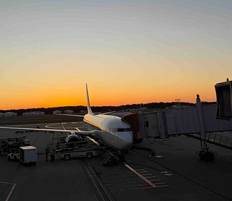 Commercial airplane parked at airport gate during sunset, connected to a jet bridge with ground crew and service vehicles nearby. Warm orange and yellow hues fill the sky, highlighting the serene yet active atmosphere of air travel operations.