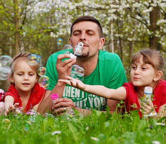Photo of a masculine-looking father figure, light-skinned man with a green tee shirt, brown hair and a goatee, blowing bubbles with two femme/girl, light-skinned children with dark blonde hair around the age of 5 and 7, wearing red tee shirts. They’re all laying in the grass behind a copse of trees.