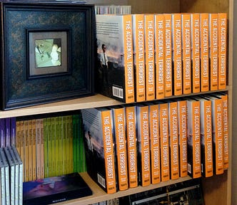A bookshelf containing various objects, including two dozen hardcover copies of The Accidental Terrorist.