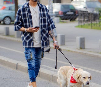 Brown-skinned man in his 30s, dreadlocks and goatie with black glasses and a white beanie, wearing white headphones, a white shirt, two necklaces, a blue and white flannel, and blue jeans with white sneakers. He’s holding a cellphone in his right hand while walking a blonde dog with a red bandana on a city street.