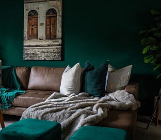 A living room with green walls and a couch with a variety of pillows in emerald green, silver, white, and gray. The room looks cozy.