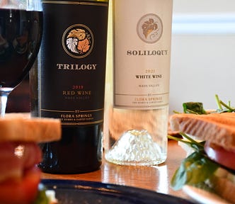 A table is set with wine and food. There is a full glass of red wine. The red wine is called Trilogy. The white wine is called Soliloquy. There is a sandwich with tomatoes, mozzarella cheese, and basil.